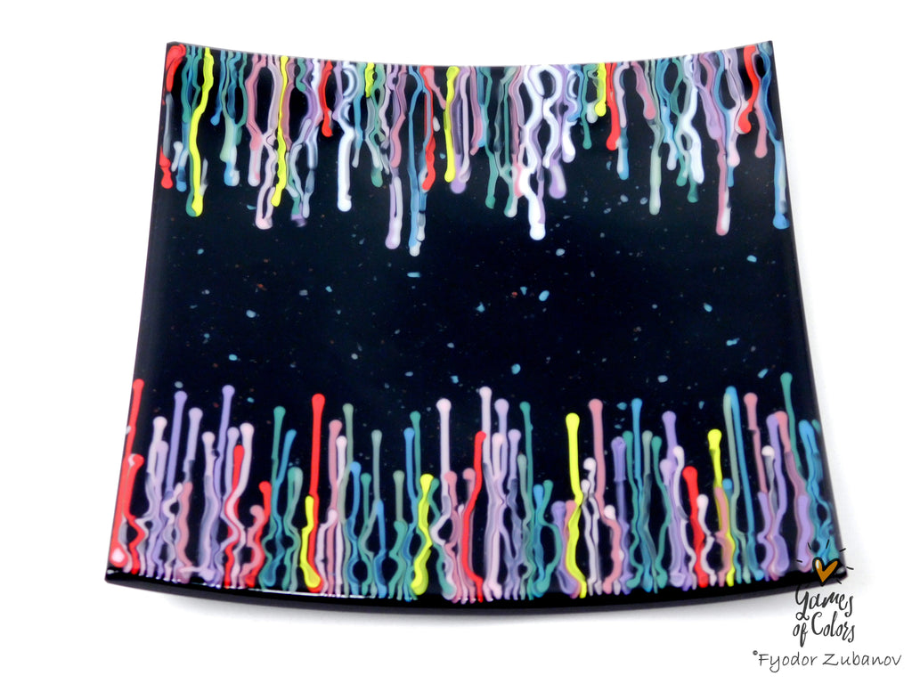 A black square glass plate featuring a colorful dripping paint pattern along the top and bottom edges, shown on a plain white background with a small artist signature in the lower right corner.