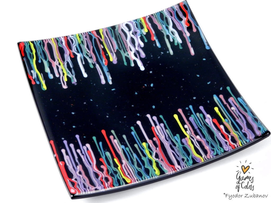A black square glass plate featuring a colorful dripping paint pattern along the top and bottom edges, shown on a plain white background with a small artist signature in the lower right corner.