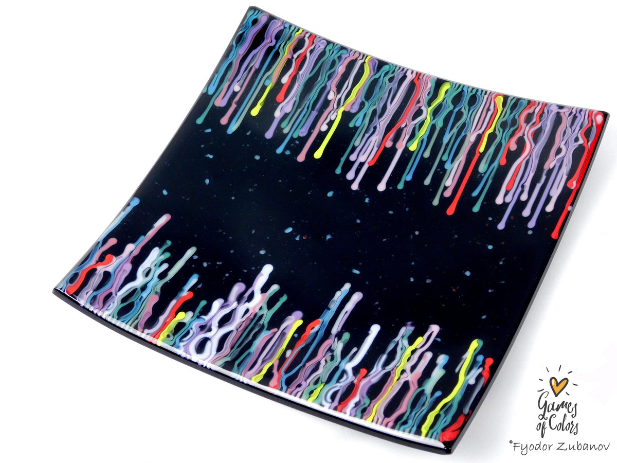 A black square glass plate featuring a colorful dripping paint pattern along the top and bottom edges, shown on a plain white background with a small artist signature in the lower right corner.
