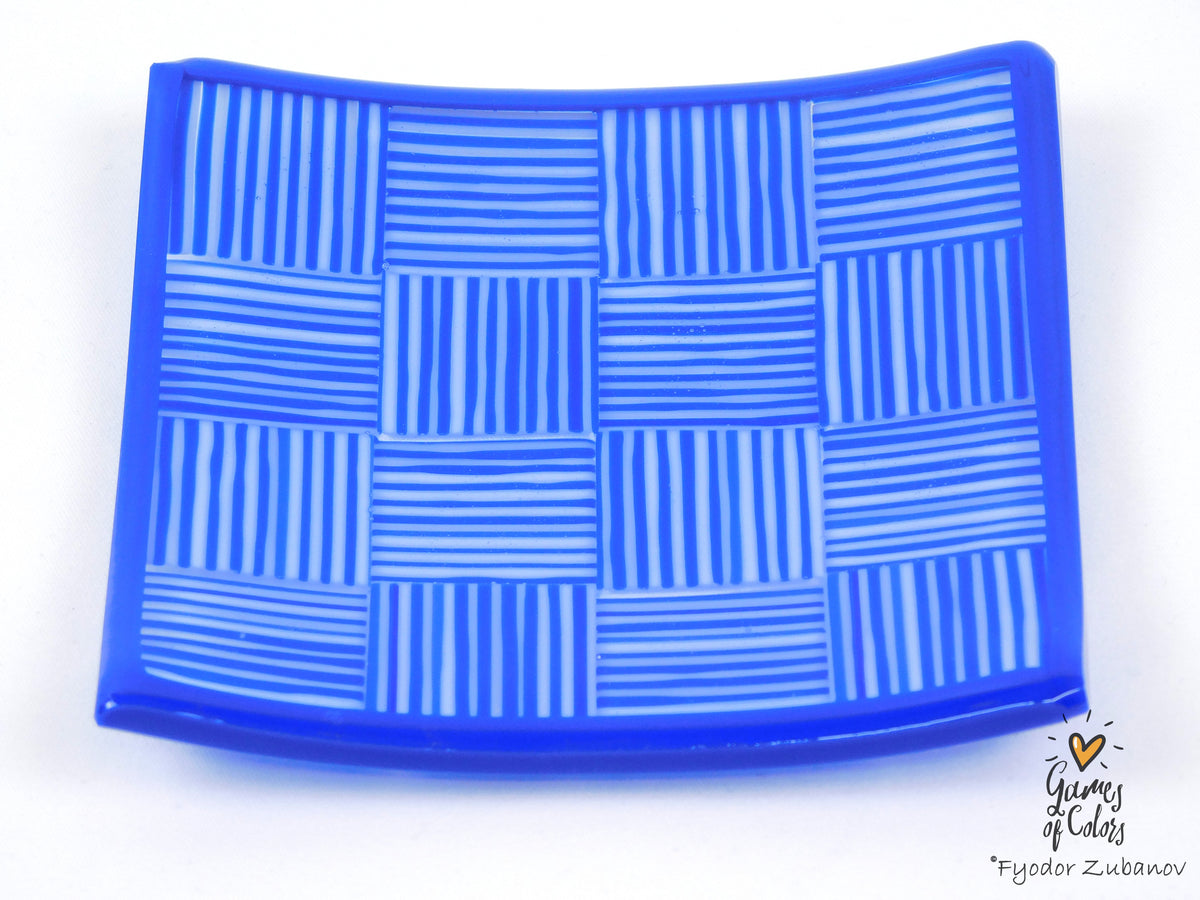 A blue square glass plate with a white woven basketweave pattern, photographed on a white background with a 'Games of Colors' artist signature in the bottom right corner.