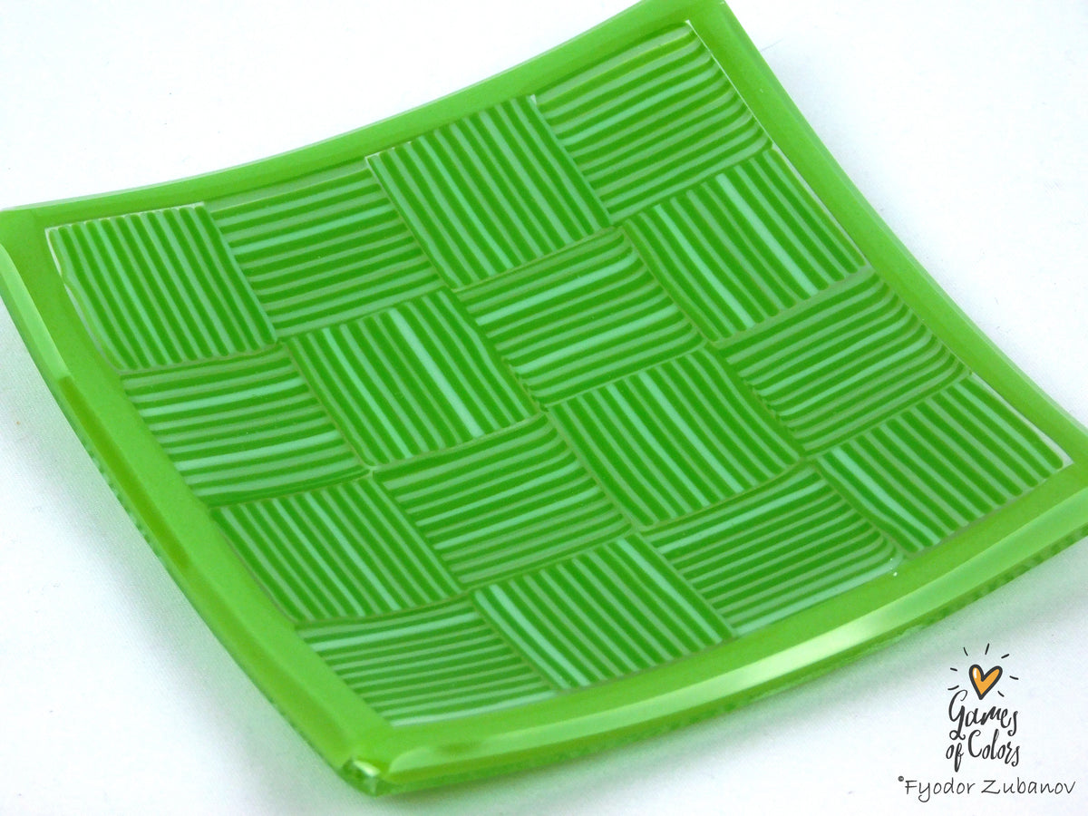 A bright green translucent square plate with a raised basketweave pattern, photographed on a plain white background. A small artist signature watermark is visible in the bottom right corner.
