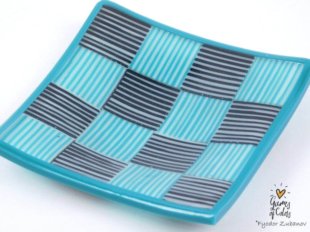 A square glass plate with a wavy edge, featuring a woven basketweave pattern of turquoise blue and dark gray striped squares, photographed on a white background with a 'Games of Colors' watermark in the lower right corner.