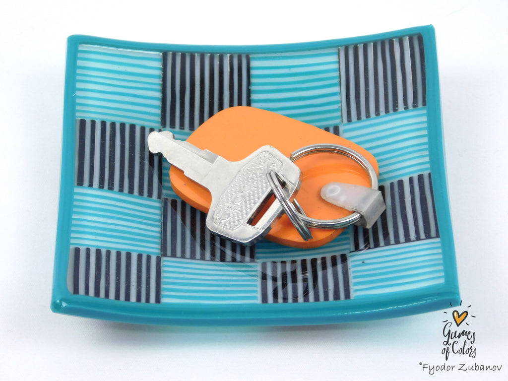 A square turquoise and black striped glass tray holding an orange keychain with silver keys, photographed on a white background.