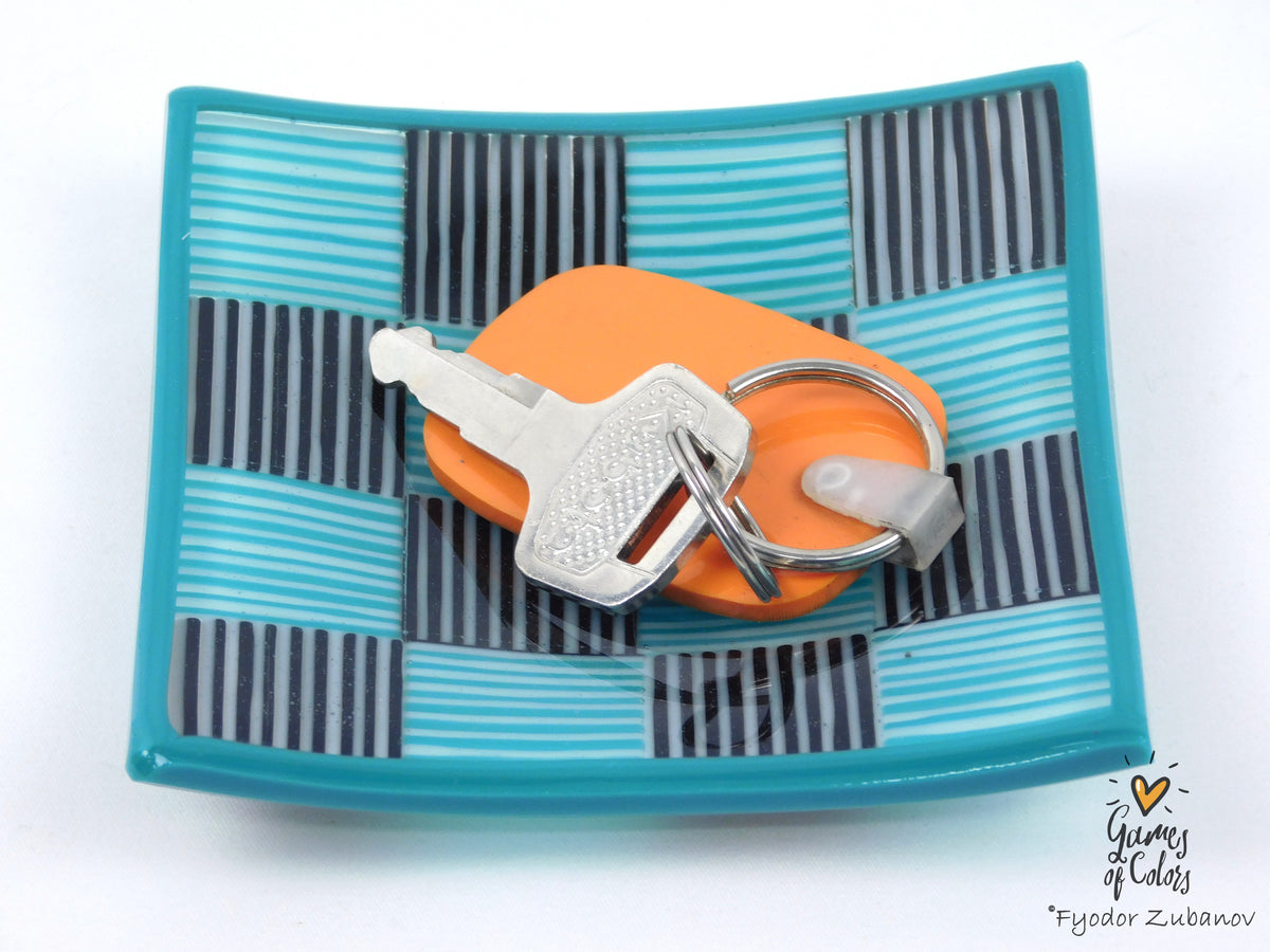A square turquoise and black striped glass tray holding an orange keychain with silver keys, photographed on a white background.