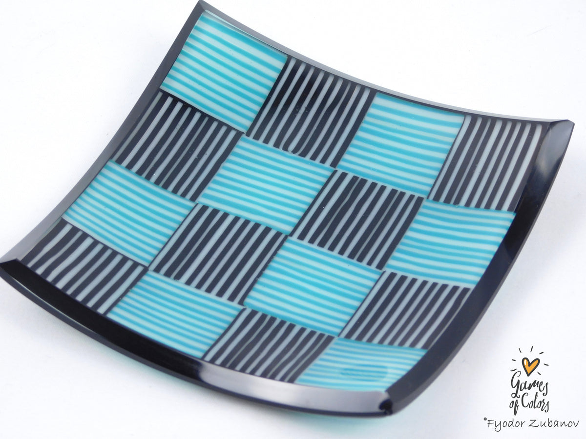 A square glass plate with a glossy finish, featuring a checkered pattern of alternating teal and black striped squares, photographed on a white background with a watermark in the bottom right corner.