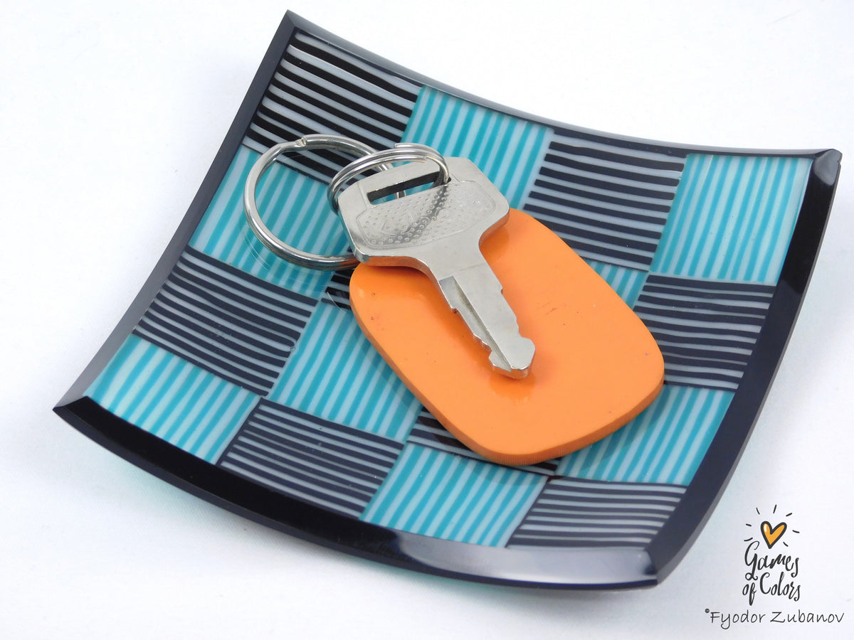 An orange keychain with a silver metal key and split ring, resting on a glossy, curved square tray with a teal, black, and white striped pattern. The photo is taken on a clean white background with a 'Games of Color' watermark in the bottom right corner.