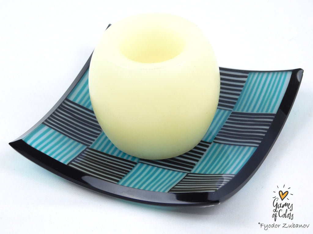 A pale yellow glass candle holder with a hollow center, displayed on a black and teal checkered glass plate against a white background.