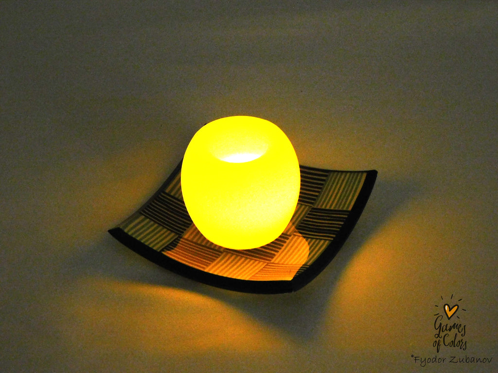 A glowing oval lamp or candle holder sits on a black square plate with a yellow and black geometric pattern, set against a dark background with warm light spill.