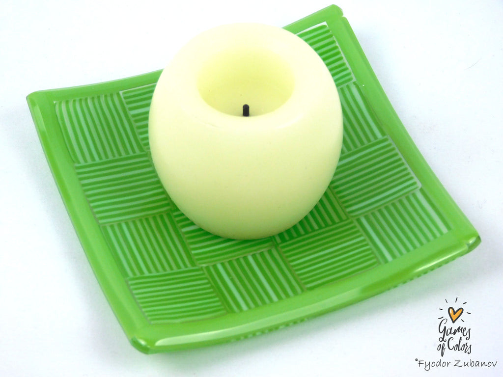 A pale yellow-green pillar candle with a black wick, displayed on a textured green square tray against a white background.