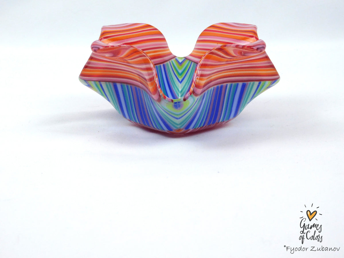 A colorful hand-blown glass bowl with wavy, scalloped edges, featuring vibrant orange, red, pink, green, blue, and purple striped patterns on a white background.