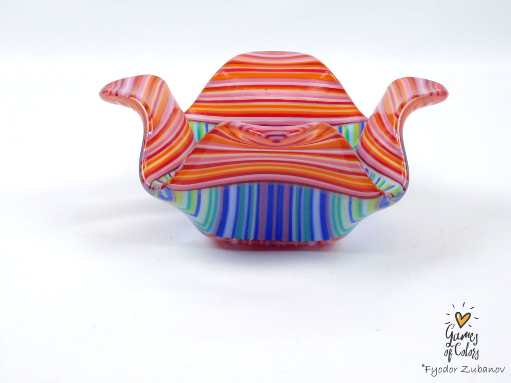 A small sculptural bowl with a colorful striped pattern, featuring red, orange, pink, yellow, green, blue, and purple bands, set against a plain white background.