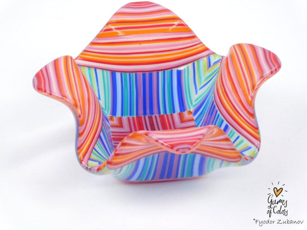 A colorful fused glass candle holder with a wavy, scalloped rim, featuring horizontal and vertical striped patterns in orange, pink, red, blue, green, and purple.