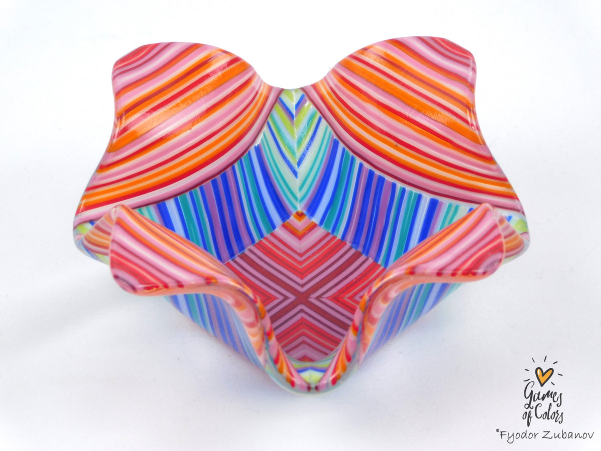 A colorful fused glass candle holder with a wavy, scalloped rim, featuring horizontal and vertical striped patterns in orange, pink, red, blue, green, and purple. 