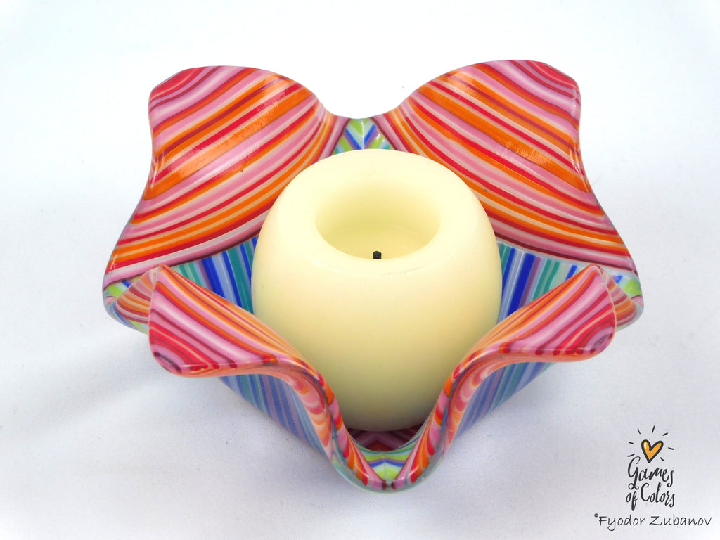 A colorful glass candle holder with a wavy, flower-like shape, featuring pink, orange, red, blue, and green striped patterns, holding a cream-colored pillar candle. The photo is taken on a plain white background with a watermark in the bottom right corner.