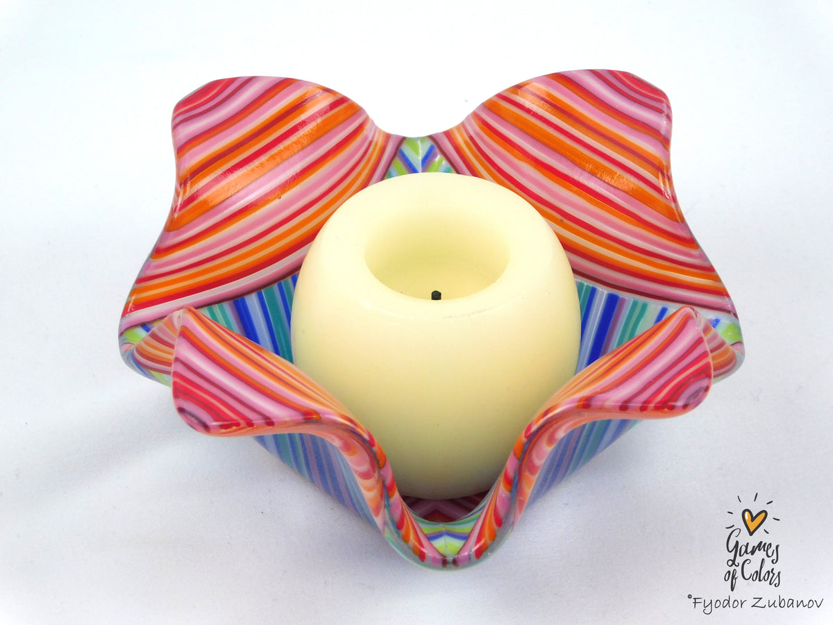 A colorful glass candle holder with a wavy, flower-like shape, featuring pink, orange, red, blue, and green striped patterns, holding a cream-colored pillar candle. The photo is taken on a plain white background with a watermark in the bottom right corner.