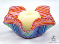 A colorful fused glass candle holder with a wavy, scalloped rim, featuring horizontal and vertical striped patterns in orange, pink, red, blue, green, and purple. A smooth, cream-colored glass candle is placed inside the holder, which is photographed on a clean white background.