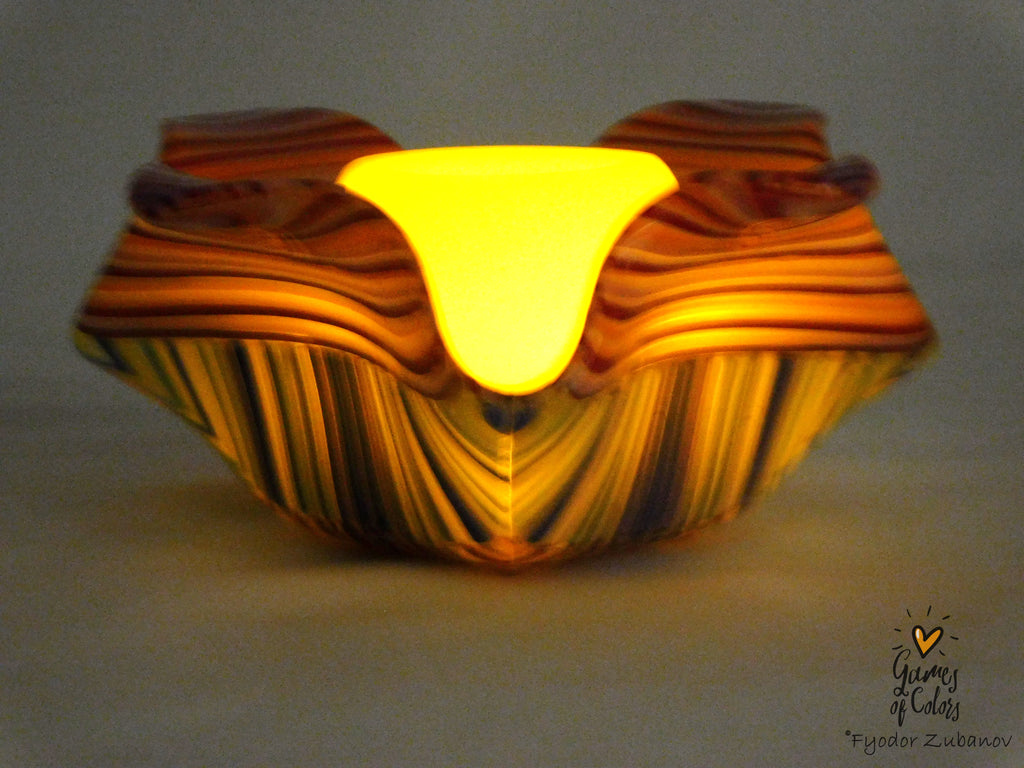 A decorative glass bowl with a wavy, layered design, featuring a glowing yellow interior and orange, yellow, and dark brown striped patterns on the exterior, set against a neutral gray background.