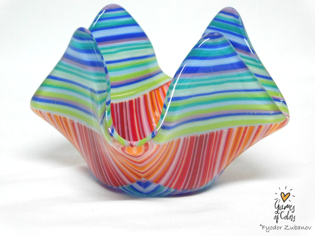 A colorful fused glass candle holder with a mountain-like flared rim, featuring horizontal striped patterns in blue, green, orange, red, and white on a white background.