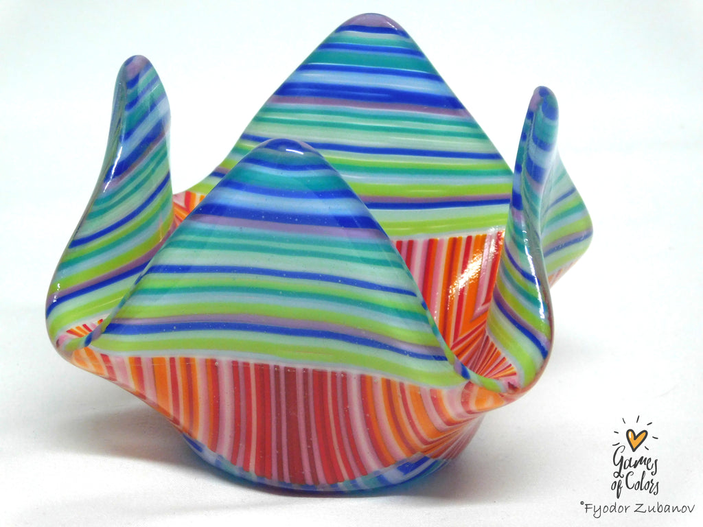 A colorful fused glass candle holder with a mountain-like flared rim, featuring horizontal striped patterns in blue, green, orange, red, and white on a white background.