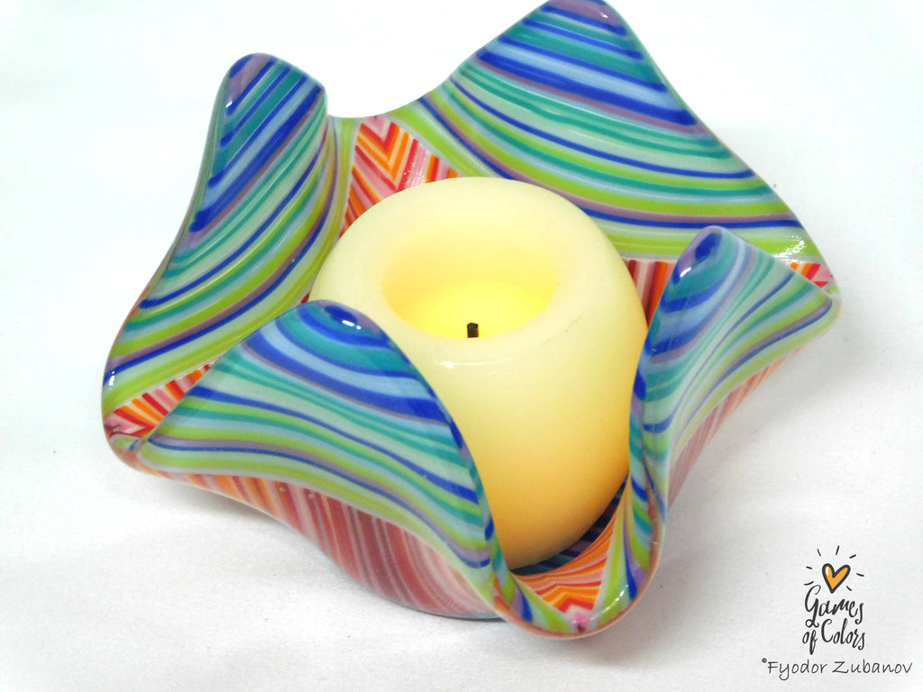 A colorful, star-shaped fused glass candle holder with wavy ridges, featuring swirls of blue, green, orange, pink, and purple. A lit cream-colored pillar candle is placed inside the holder, which is set against a plain white background.