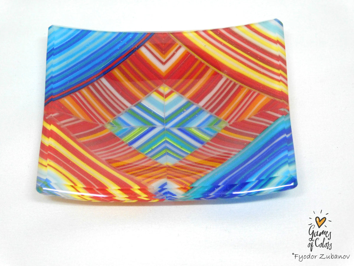 A square glass plate with a vibrant, colorful geometric pattern featuring red, orange, yellow, blue, and green stripes and diamond shapes, photographed on a white background.