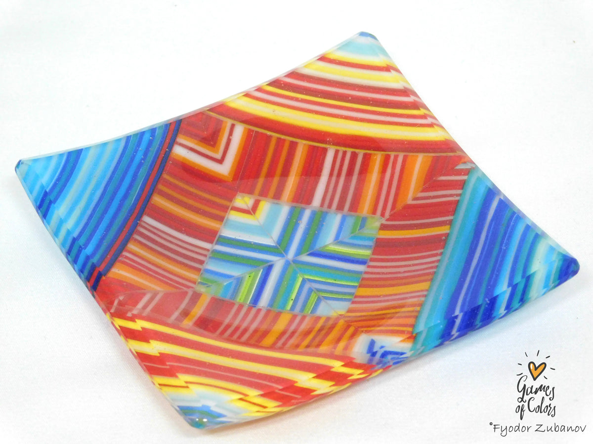 A square fused glass plate with a vibrant multicolor geometric pattern, featuring stripes and concentric squares in red, orange, yellow, blue, green, and white, photographed on a white background.