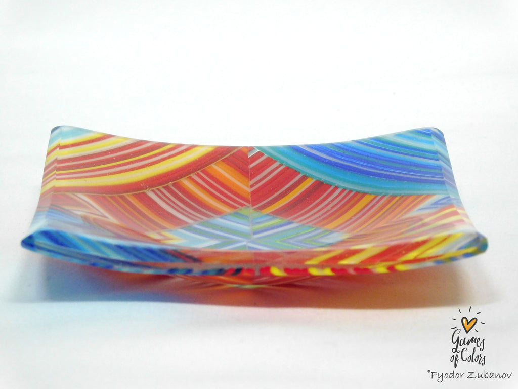 A square, shallow glass dish with a vibrant, multicolored swirl pattern of red, orange, yellow, blue, and green, photographed on a white background with a soft shadow.