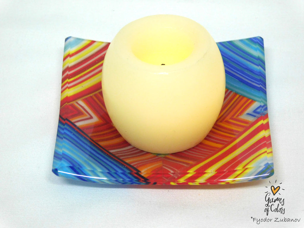 A cream-colored glass candle holder with a wavy, colorful geometric pattern in red, orange, yellow, and blue, featuring a lit cream candle inside, set against a plain white background.