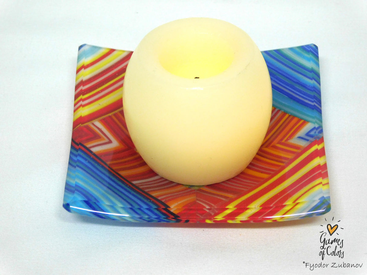 A cream-colored glass candle holder with a wavy, colorful geometric pattern in red, orange, yellow, and blue, featuring a lit cream candle inside, set against a plain white background.