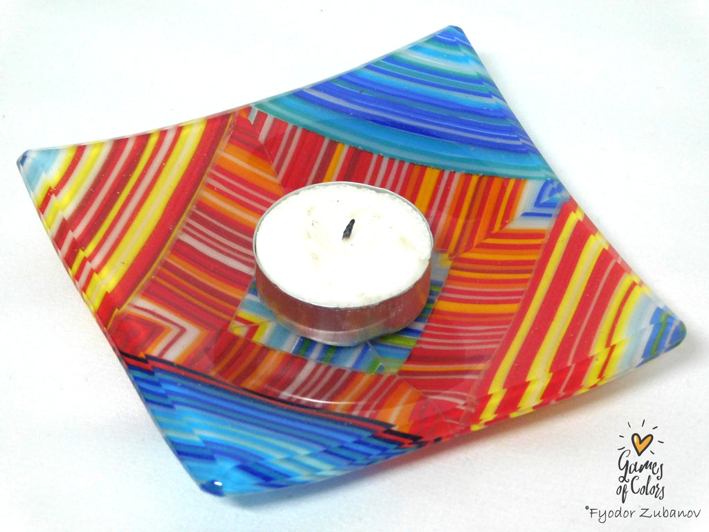 A square glass dish with a vibrant multicolor abstract pattern of red, orange, yellow, blue, and white stripes, holding a small white tea light candle on a white background.