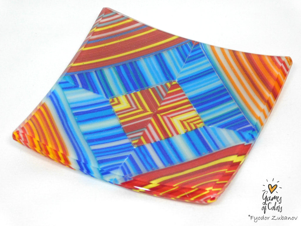 A square glass plate with a colorful geometric pattern featuring blue, red, orange, yellow, and white stripes, photographed on a white background with a watermark in the bottom right corner.