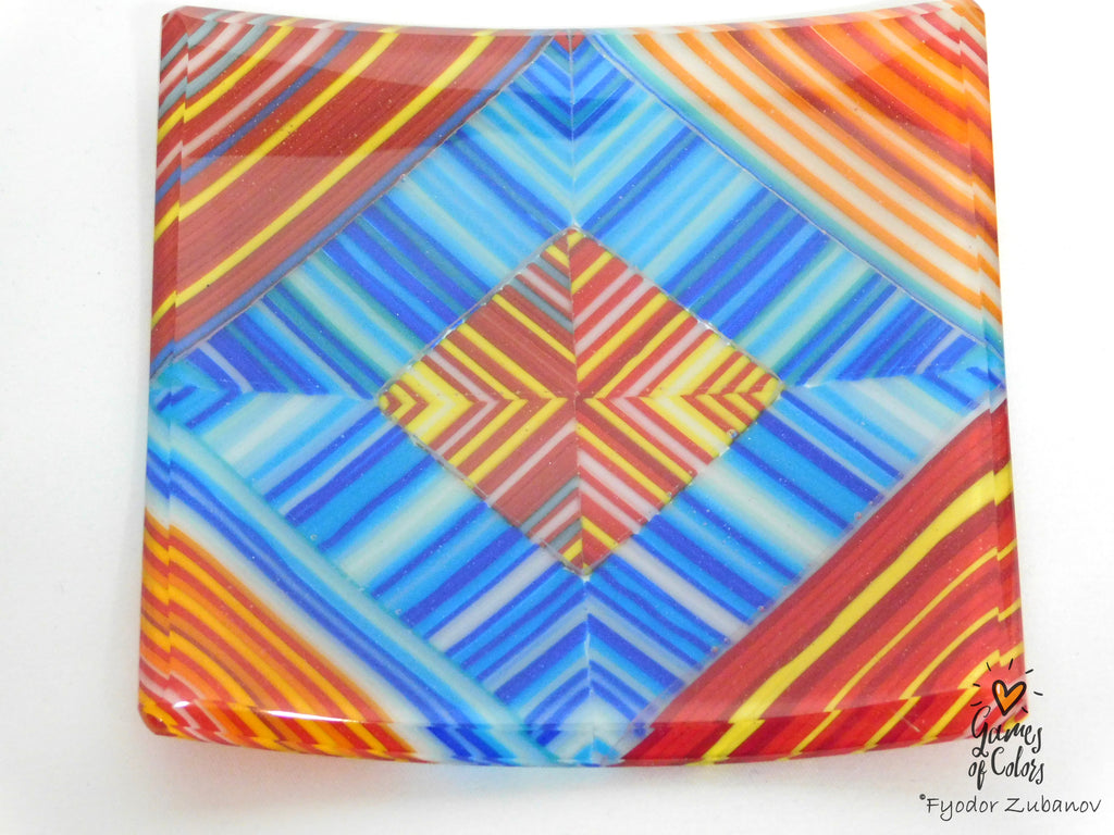 A square glass plate featuring a vibrant geometric pattern with concentric diamond shapes and radiating stripes in blue, red, orange, yellow, and white, photographed on a plain white background.
