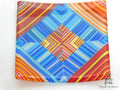 A square glass plate featuring a vibrant geometric pattern with concentric diamond shapes and radiating stripes in blue, red, orange, yellow, and white, photographed on a plain white background.