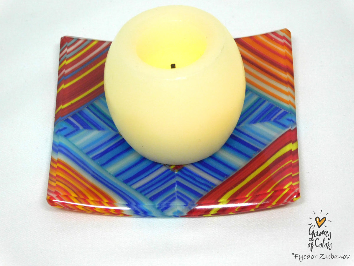 A glass candle holder with a colorful geometric pattern in blue, red, orange, and yellow, holding a lit cream-colored glass candle, set against a plain white background.