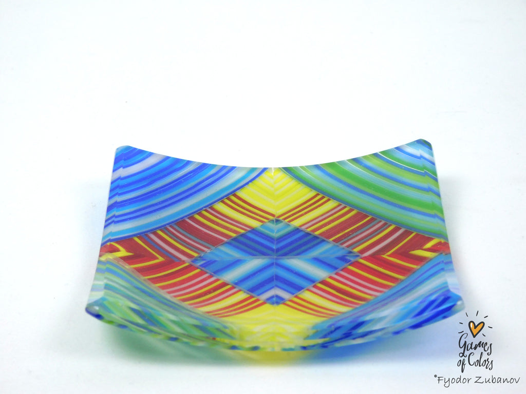 A square glass plate with a vibrant, symmetrical geometric pattern featuring blue, yellow, green, red, and orange stripes, photographed on a plain white background.
