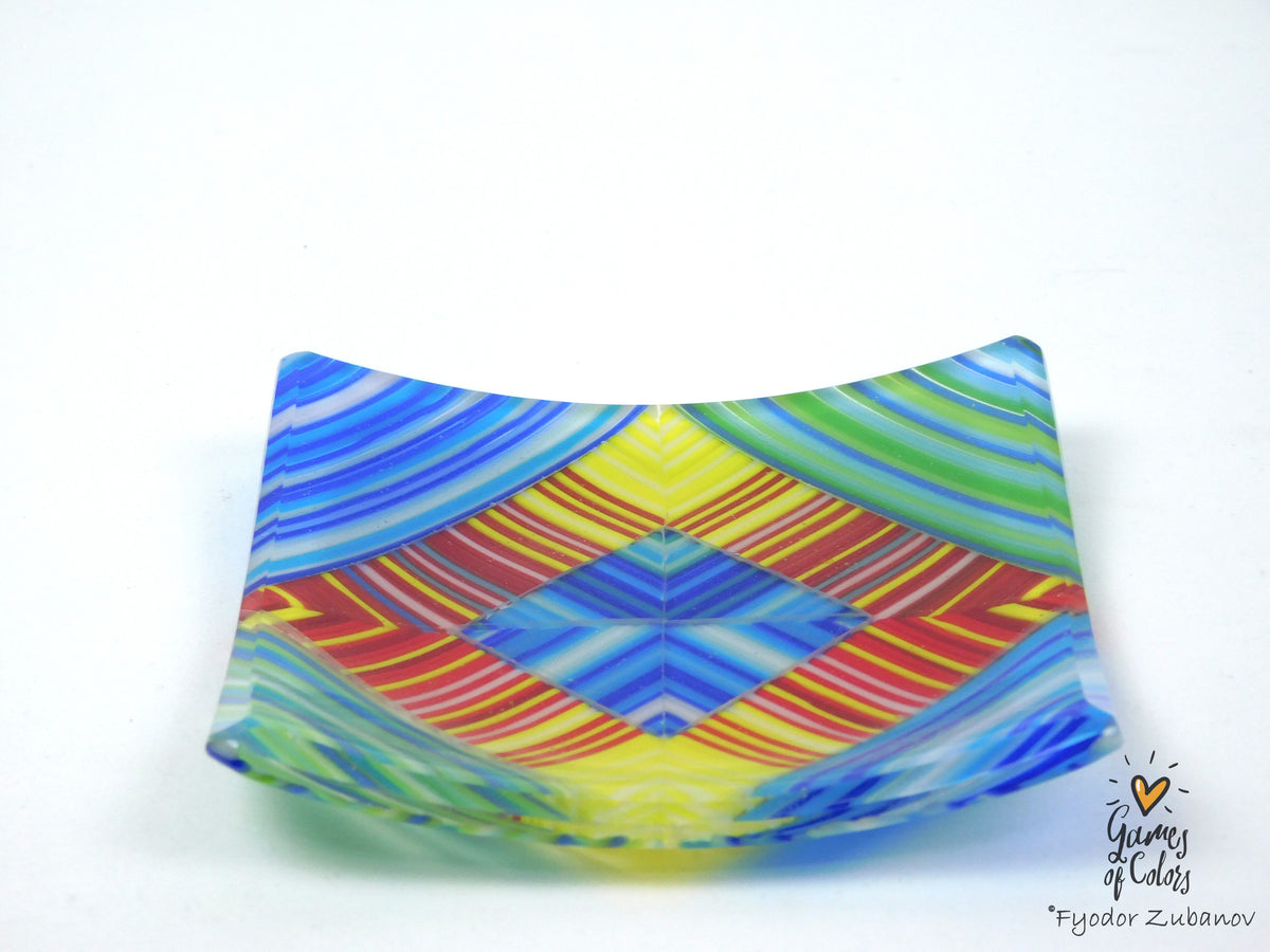 A square glass plate with a vibrant, symmetrical geometric pattern featuring blue, yellow, green, red, and orange stripes, photographed on a plain white background.