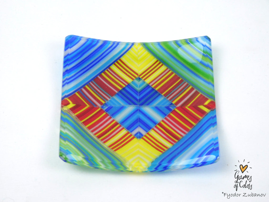 A square glass plate with a vibrant, multicolored geometric pattern featuring blue, yellow, red, green, and orange stripes, photographed on a white background.