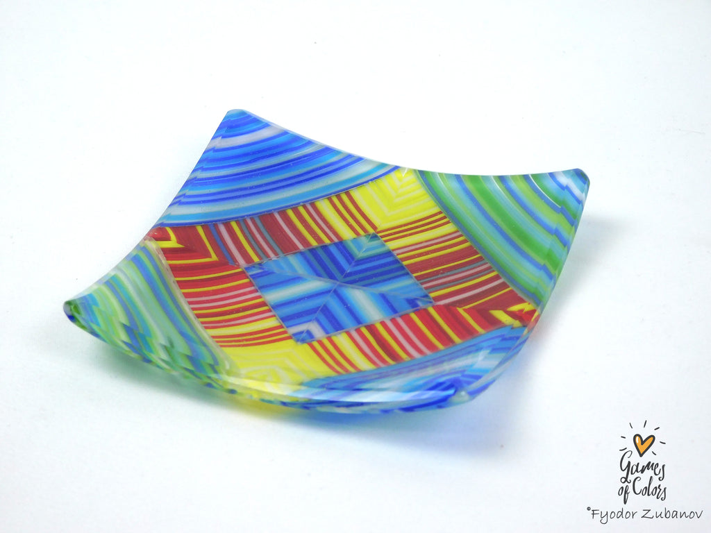 A handmade, asymmetrical square glass plate with a vibrant, multicolored geometric pattern featuring blue, yellow, green, red, and orange stripes, shown on a plain white background.