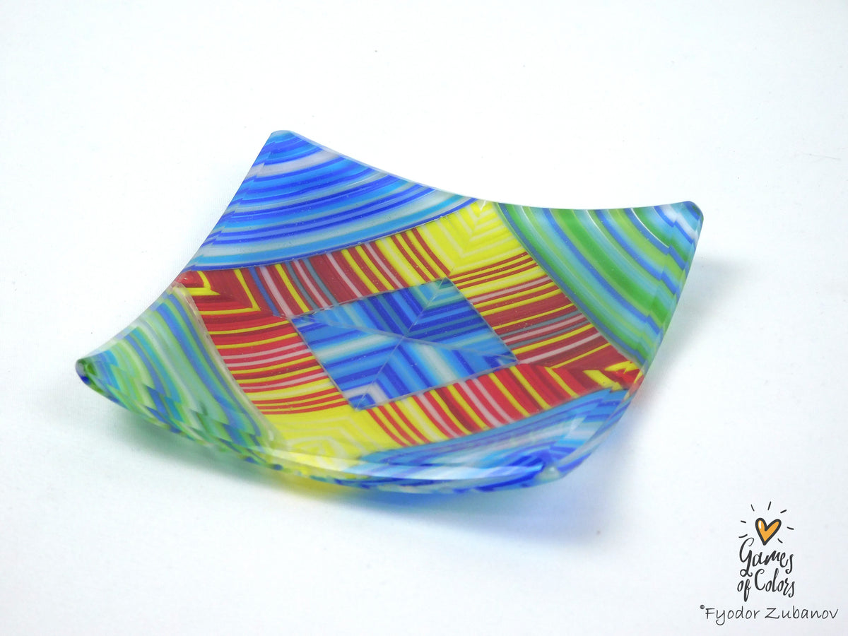 A handmade, asymmetrical square glass plate with a vibrant, multicolored geometric pattern featuring blue, yellow, green, red, and orange stripes, shown on a plain white background.