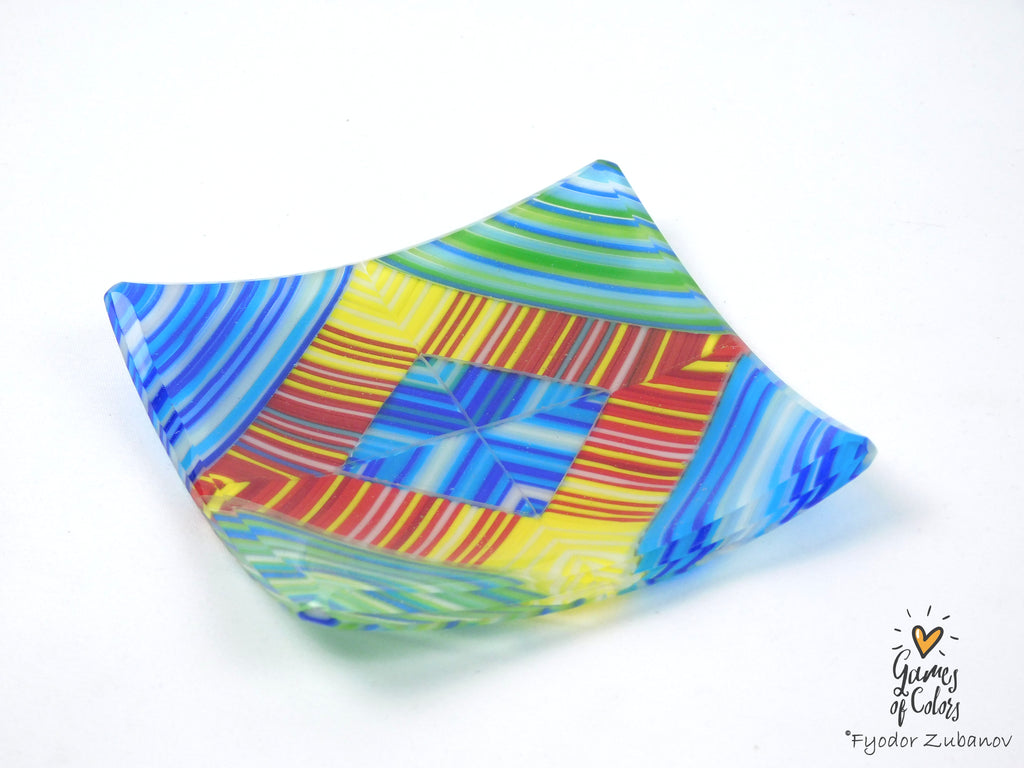 A colorful, fused glass dish with a geometric pattern of blue, yellow, green, and red stripes, photographed on a white background with a watermark in the bottom right corner.