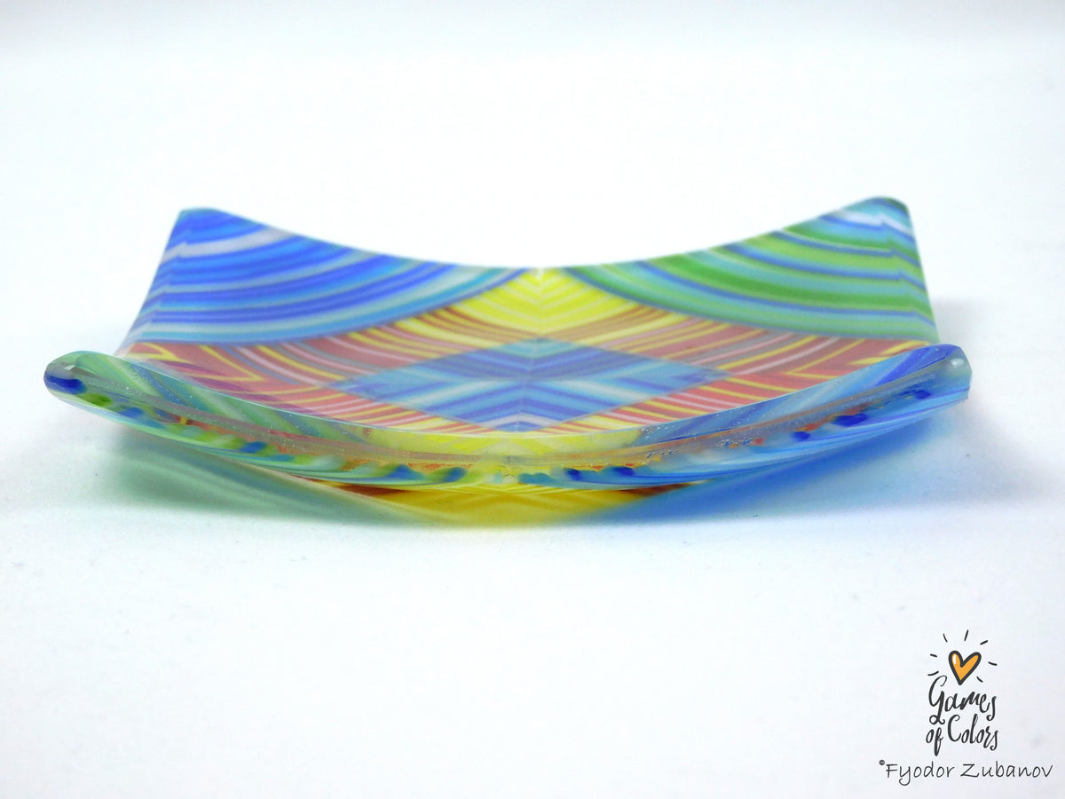 A handmade, translucent glass plate with a colorful, wavy pattern of blue, green, yellow, pink, and orange swirls, photographed on a white background.