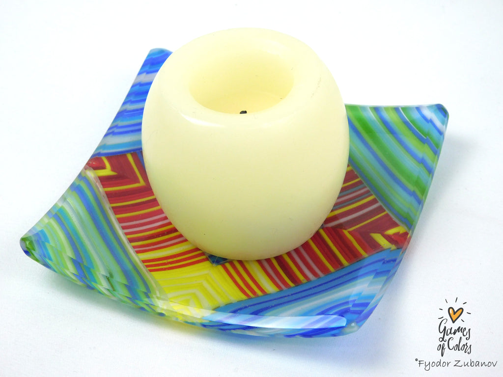 A cream-colored pillar candle displayed in a square, colorful glass dish with blue, green, yellow, and red patterns, on a plain white background.