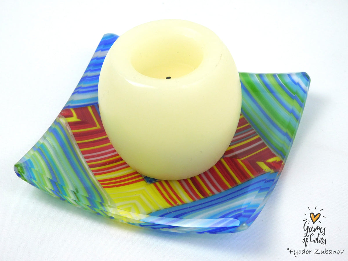 A cream-colored pillar candle displayed in a square, colorful glass dish with blue, green, yellow, and red patterns, on a plain white background.