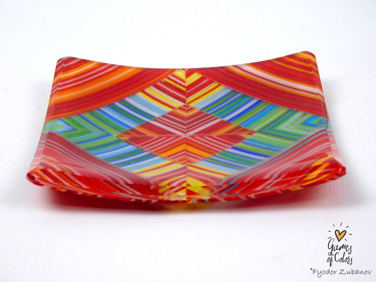 A square glass plate with a vibrant, multicolored geometric pattern featuring red, orange, yellow, green, and blue stripes, photographed on a white background.
