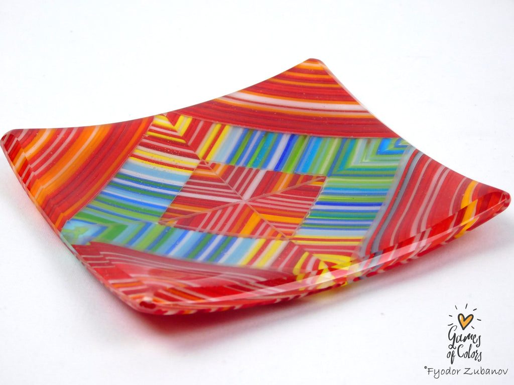 A square glass dish with a vibrant multicolor geometric pattern featuring red, orange, yellow, green, blue, and white stripes, photographed on a white background with a watermark in the bottom right corner.