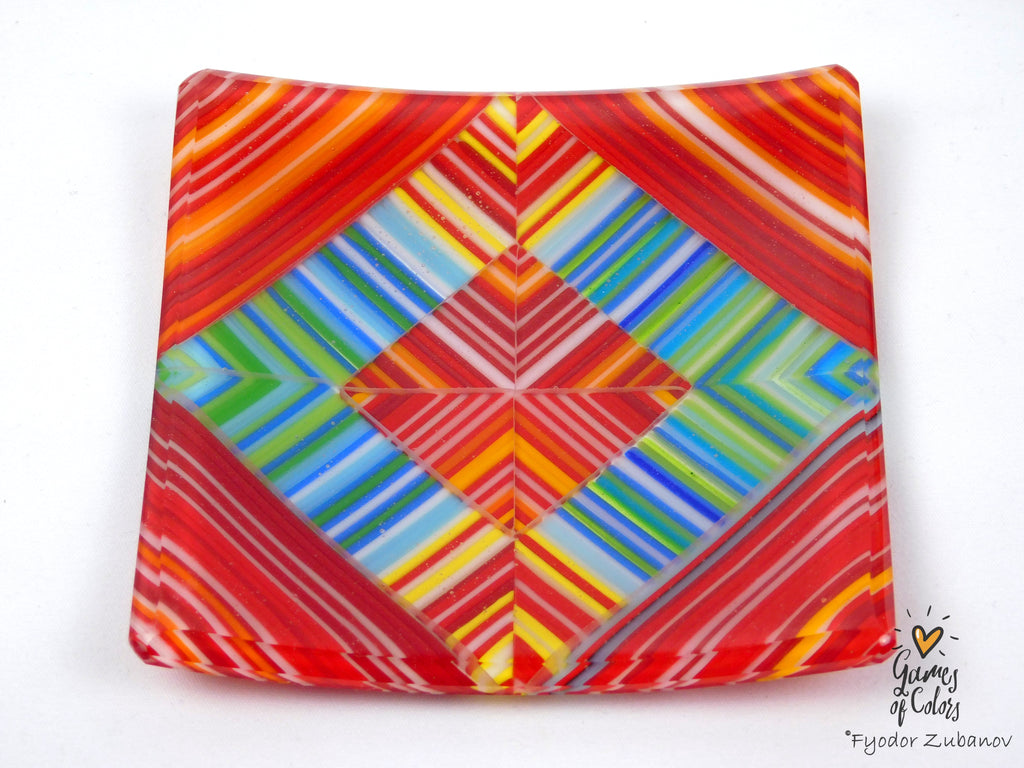 A square glass plate featuring a vibrant, symmetrical geometric pattern with stripes of red, orange, yellow, green, blue, and purple on a white background.