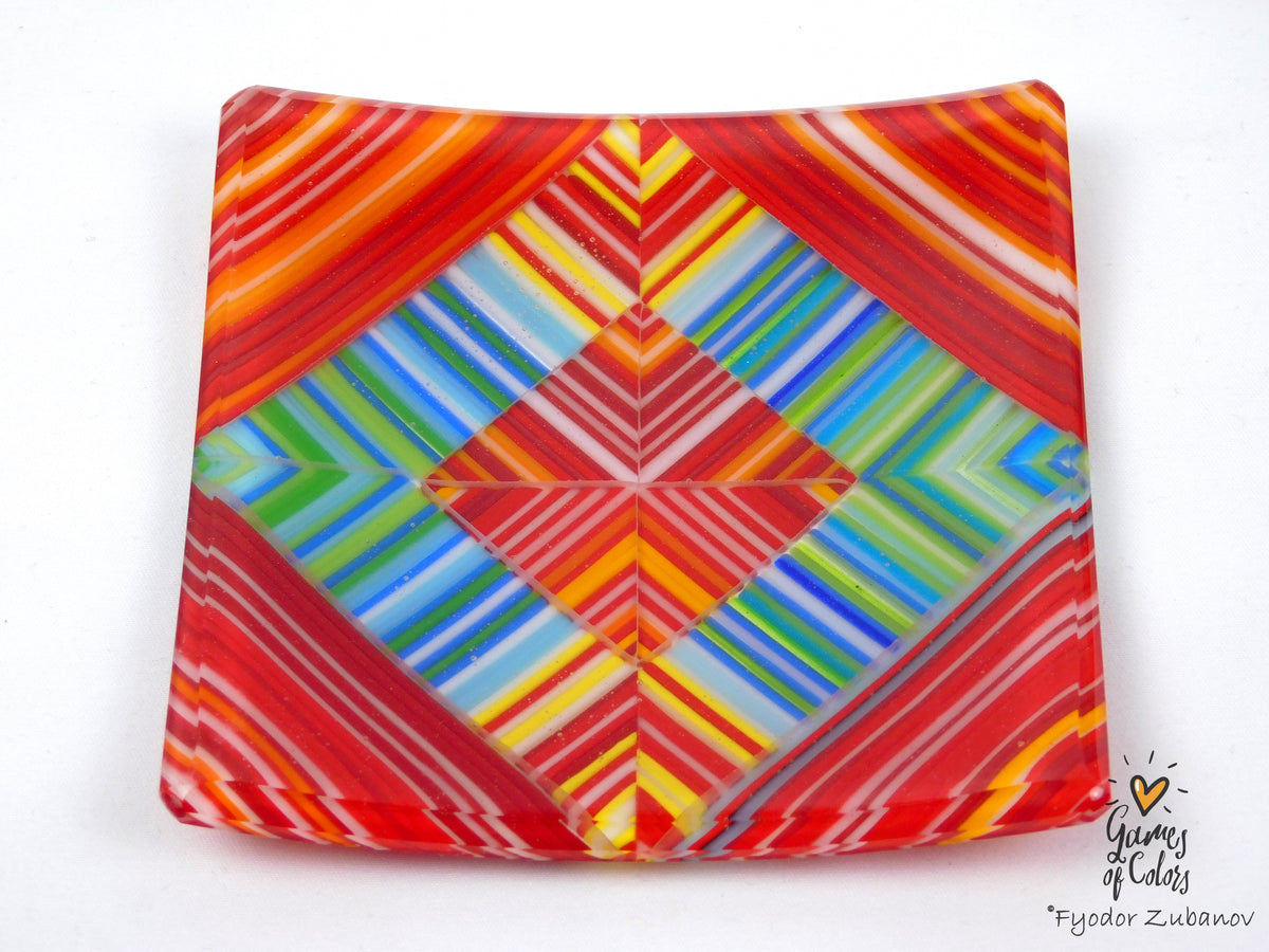 A square glass plate featuring a vibrant, symmetrical geometric pattern with stripes of red, orange, yellow, green, blue, and purple on a white background.