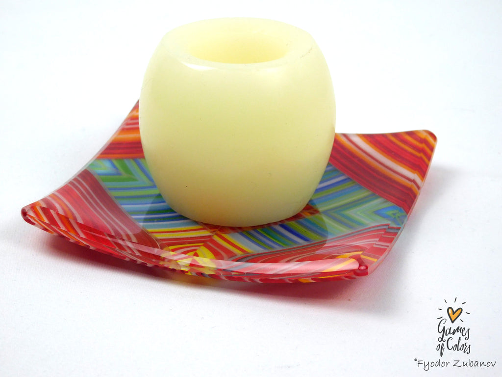 A pale candle with a rounded, hollow center, displayed on a square, colorful glass plate with red, orange, yellow, blue, and green stripes against a white background.