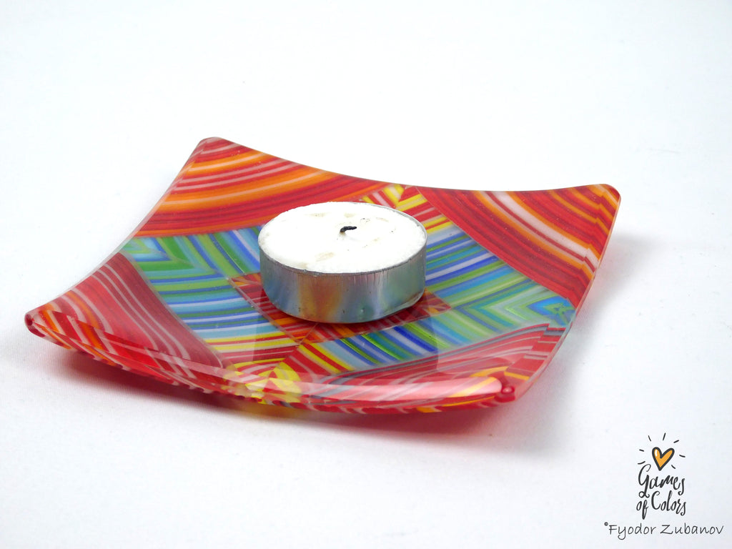 A square glass candle holder with a vibrant multicolor swirl pattern of red, orange, yellow, green, and blue, holding a small silver tea light candle, set against a plain white background.