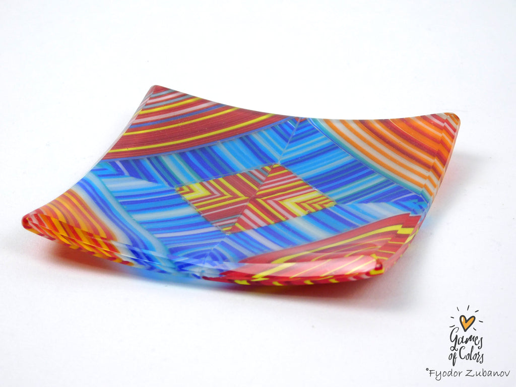 A square, fused glass plate with a vibrant multicolor geometric pattern of blue, red, orange, yellow, and white stripes, shown on a plain white background.